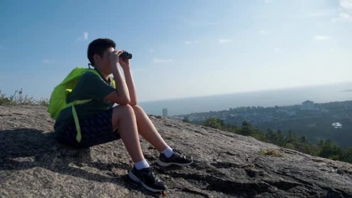 快乐的亚洲背包客男孩，早上坐在山顶上，用双筒望远镜看着风景，是家庭徒步旅行的概念。