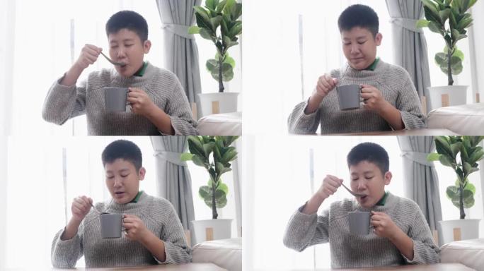 亚洲男孩穿着灰色毛衣，在杯子里喝热巧克力，在家坐在窗户附近，生活方式概念。
