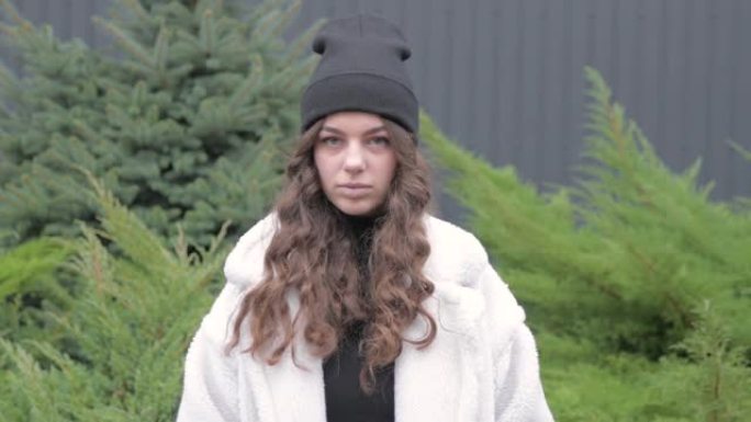 看起来严肃的年轻欧洲女性漂亮的脸看着相机独自站在户外摆姿势。画像前。穿着暖和的衣服，夹克和帽子在院子