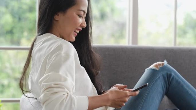 亚洲女人在手机上与朋友聊天，开心的时刻，坐在家里的沙发上。社交媒体生活方式