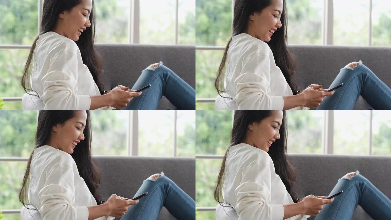 亚洲女人在手机上与朋友聊天，开心的时刻，坐在家里的沙发上。社交媒体生活方式