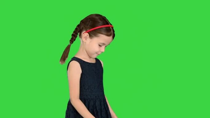 穿着黑色连衣裙的辫子可爱的小女孩低头看着她的脚，然后在绿色屏幕上对着镜头微笑，色键
