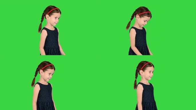 穿着黑色连衣裙的辫子可爱的小女孩低头看着她的脚，然后在绿色屏幕上对着镜头微笑，色键