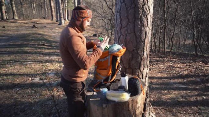 主题徒步旅行。一名高加索旅游男子打开一个橙色背包，拿出他的东西，放在森林的树桩上。露营的设备和东西。