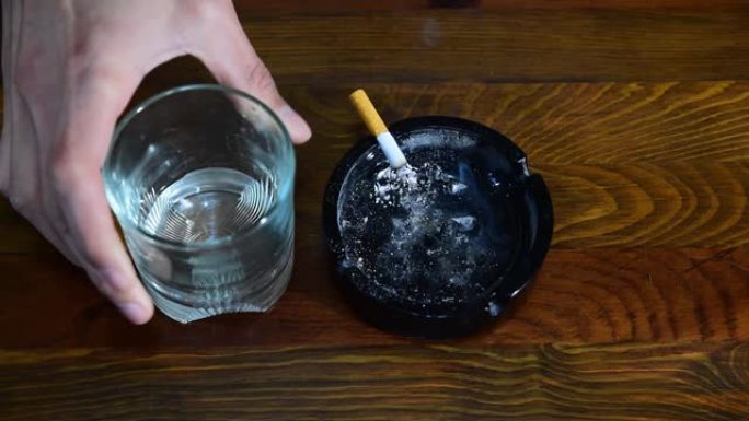 肮脏的陶瓷烟灰缸，上面燃烧着香烟，在木条桌子上抽烟，一杯酒精饮料被沮丧的酒精和吸烟者从上面看