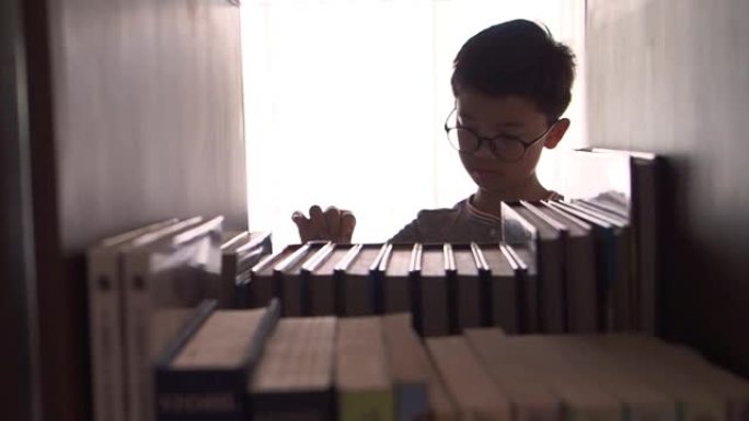 亚洲男孩在家看书，书架上的书前景模糊，相机朝右侧平移，背景明亮。