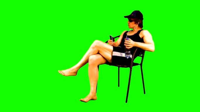 有魅力的运动员坐在扶手椅上，晒日光浴，看着他的智能手机，在上面听音乐，享受和喝色度键的瓶子。