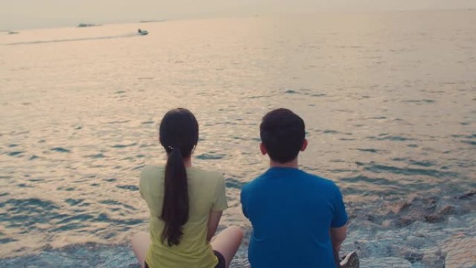 亚洲年轻夫妇跑步后休息，坐在海边看着美丽的日落。健康生活方式的概念。