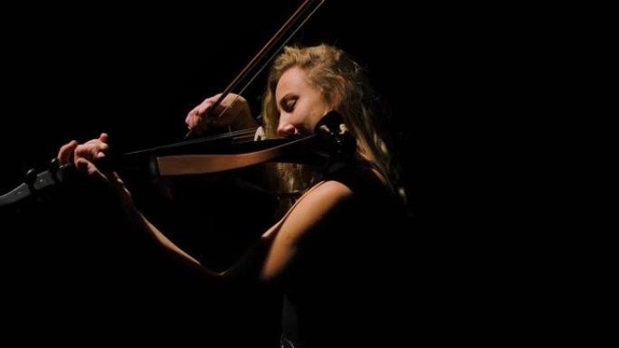 一个迷人的年轻女子在黑暗中拉小提琴的侧视图。小提琴手在黑色背景下用录音棚灯光表演古典独奏会。特写。慢