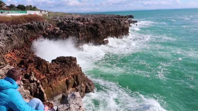 强大的海浪撞击岩石。纯粹的力量，自然力量的展示。强能量。对抗地球的海洋力量。年轻人坐在海边的悬崖上，