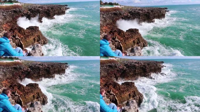 强大的海浪撞击岩石。纯粹的力量，自然力量的展示。强能量。对抗地球的海洋力量。年轻人坐在海边的悬崖上，