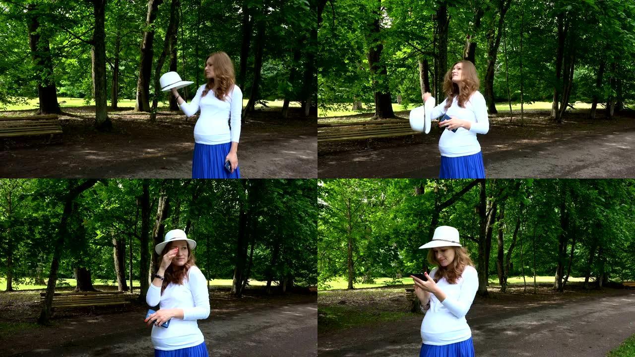 迷人的孕妇戴着帽子和电话在公园散步