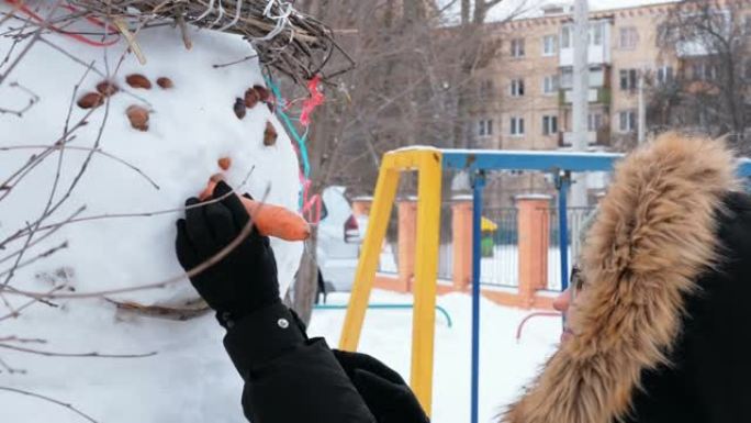 一个戴着绿色帽子和蓬松兜帽的迷人女人为雪人准备了胡萝卜