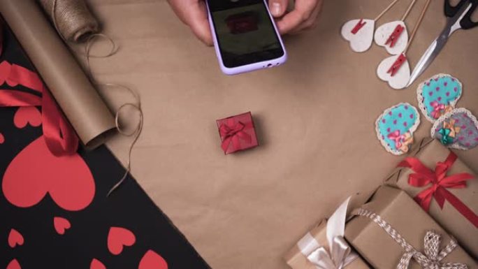 男性双手拿着电话，并为一个红色的小盒子拍照。包装纸和红心。情人节礼物概念