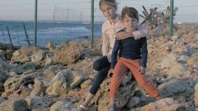 两个可怜的孤儿难民，男孩和女孩坐在靠近海边的石头上，后面是移民用的铁丝网