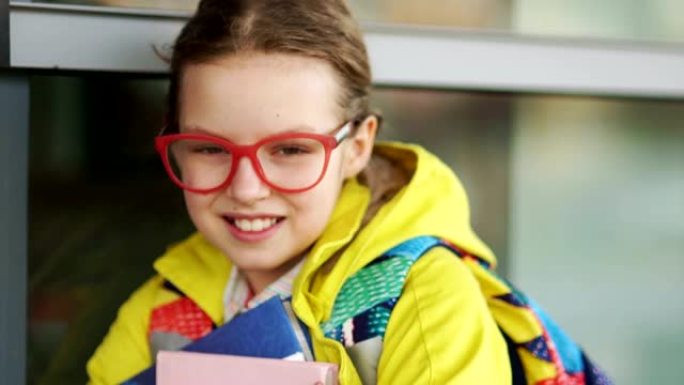 十岁女学生对着镜头开心地微笑。戴着红色眼镜和黄色外套的女孩手里拿着教科书
