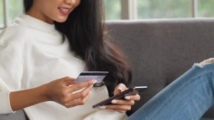 年轻的亚洲女性使用智能手机和信用卡支付在线购物，坐在家里的沙发上。数字时代的生活方式