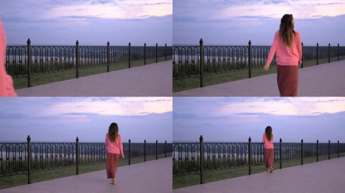 女孩沿着栅栏和夕阳散步。黑发女人把手放在栅栏上