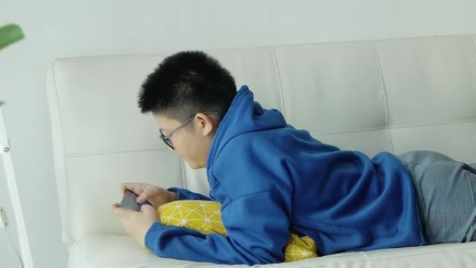 亚洲男孩戴着眼镜玩智能手机在线游戏，躺在家里的沙发上，生活方式概念。