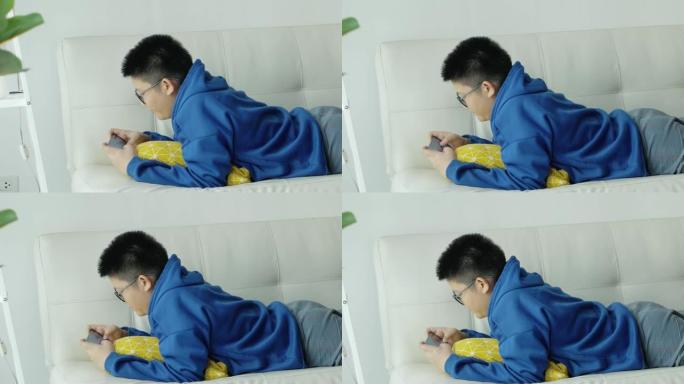 亚洲男孩戴着眼镜玩智能手机在线游戏，躺在家里的沙发上，生活方式概念。