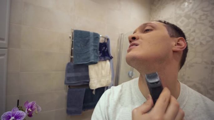 男性美容，漂亮的男人用电动修剪器在浴室的镜子前剃毛胡茬