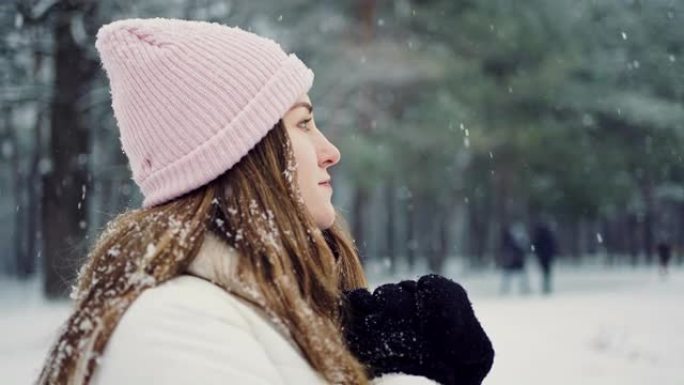 特写镜头，年轻女子站在公园的降雪下，用手套加热双手，慢动作