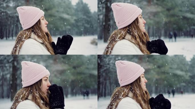 特写镜头，年轻女子站在公园的降雪下，用手套加热双手，慢动作