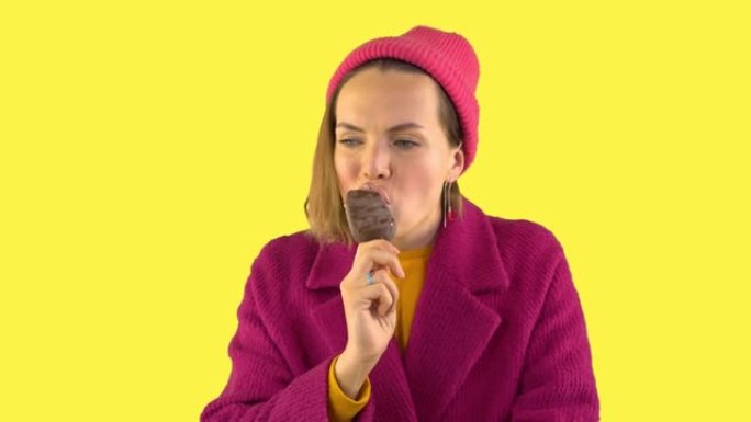 女人在巧克力釉的棍子上咬冰淇淋，然后冷冻，她变冷了。女人穿着针织帽，黄色背景上的红色外套在工作室