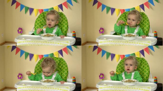 饥饿的女婴拿着勺子坐在喂食椅上，从盘子里吃粥