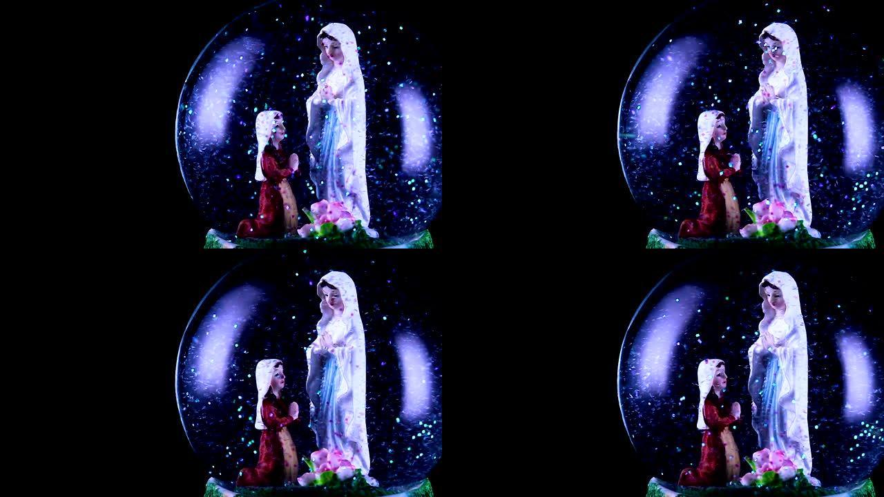 圣母玛利亚和一个信徒跪在雪球里。背景完全是黑色的，视频右侧是雪球。雪球里有很多闪闪发光的闪光。