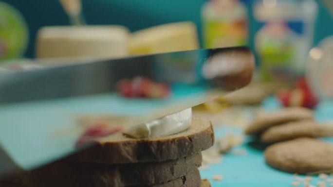 特写柔和的黄油或酸奶油在蓝色背景的桌子上用金属刀在乡村黑麦面包上慢动作。特写制作酸奶油三明治。用相机