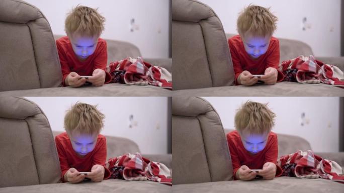 小男孩躺在沙发上，手里拿着智能手机，玩手机游戏或使用社交网络。儿童从小就依赖小工具和互联网的现代问题