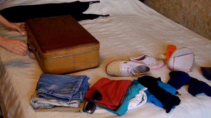 一个女人把一个棕色的老式手提箱放在床上，然后打开它。床上的衣服。快乐的trevels，度假概念。