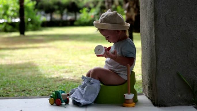 可爱的蹒跚学步的男孩，坐在度假胜地露台后门廊的便盆上，玩玩具