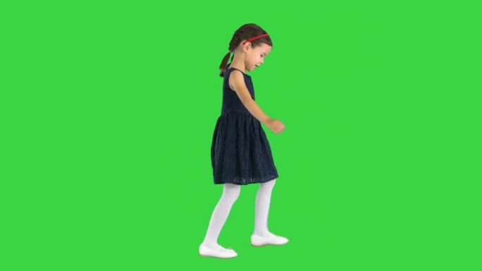 穿着黑色连衣裙的辫子可爱的小女孩低头看着她的脚，然后在绿色屏幕上微笑，色键