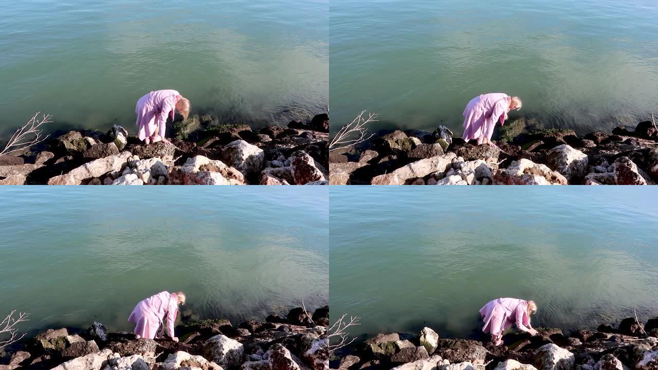 穿着粉红色外套和靴子的年轻金发女子在阳光明媚的天气里走过大海石，试图不掉入水中。滑石头，不舒服的衣服