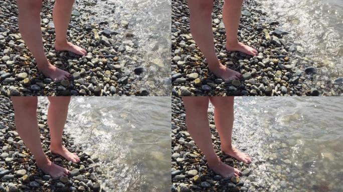 夏季晴天，女性的脚被温暖的海浪冲刷。概念。美丽的黑色指甲女性脚站在卵石滩的俯视图