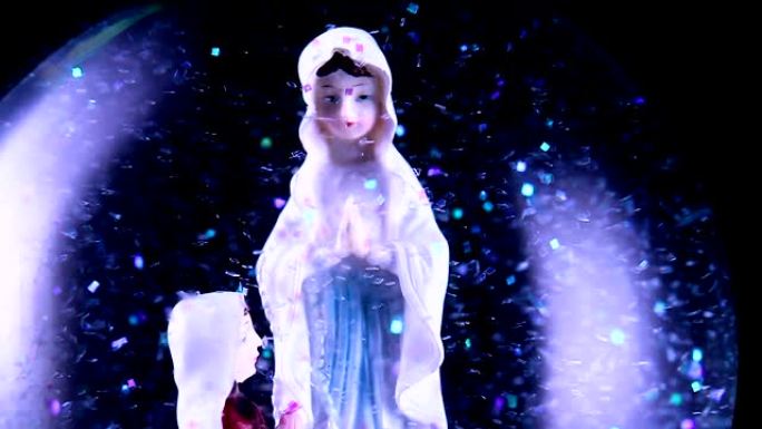 圣母玛利亚和一个信徒跪在雪球里的特写镜头。背景完全是黑色的。雪球里有很多闪闪发光的闪光。