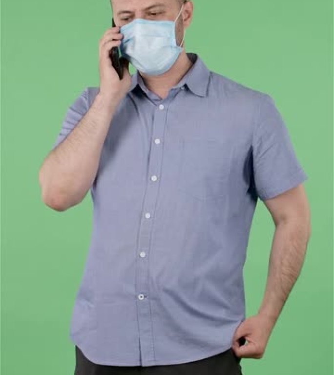 戴着医用口罩的中年男子在智能手机上聊天的肖像。穿着蓝色衬衫的秃头男性在工作室的绿色屏幕上摆姿势。慢动