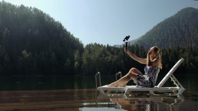 女人在智能手机的手持电影万向节稳定拍摄。女孩躺在码头的日光浴床上自拍。博客播放视频博客。