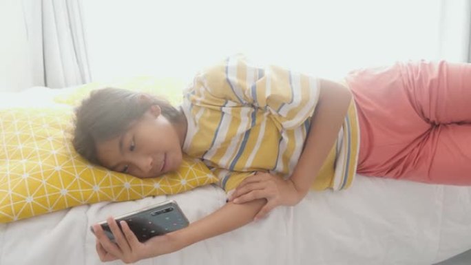 快乐的亚洲女孩躺在床上，用智能手机在家里有阳光的窗户附近看电影或病毒视频，生活方式概念。