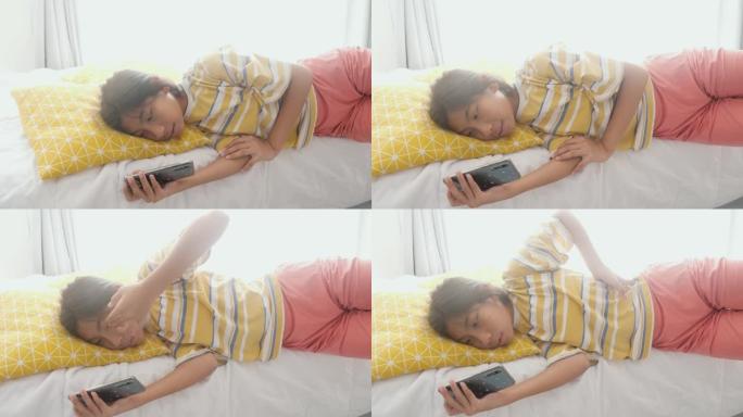 快乐的亚洲女孩躺在床上，用智能手机在家里有阳光的窗户附近看电影或病毒视频，生活方式概念。