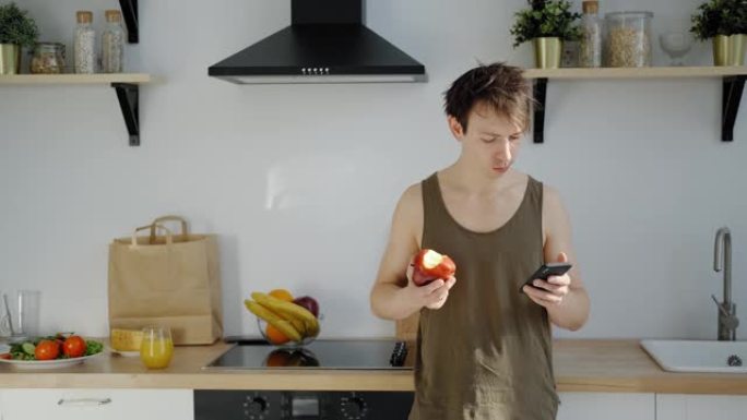 一个穿着无袖t恤的男人吃着一个红苹果，看着智能手机