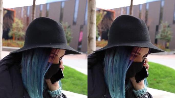 蓝色头发戴着黑色防护面具，戴着帽子在外面打电话的学生女孩。垂直视频