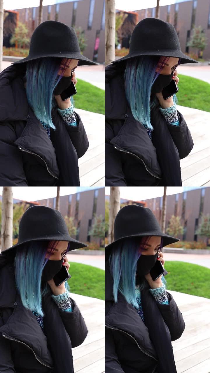 蓝色头发戴着黑色防护面具，戴着帽子在外面打电话的学生女孩。垂直视频