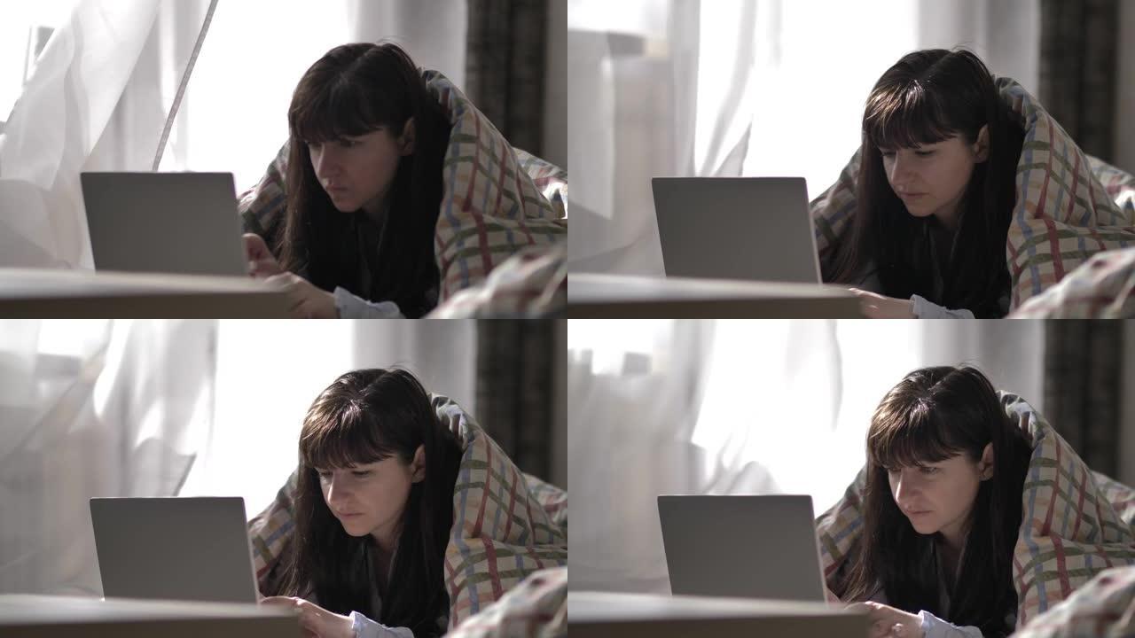 年轻女子躺在床上，上面铺着从窗户吹来的毯子，在笔记本电脑上打字