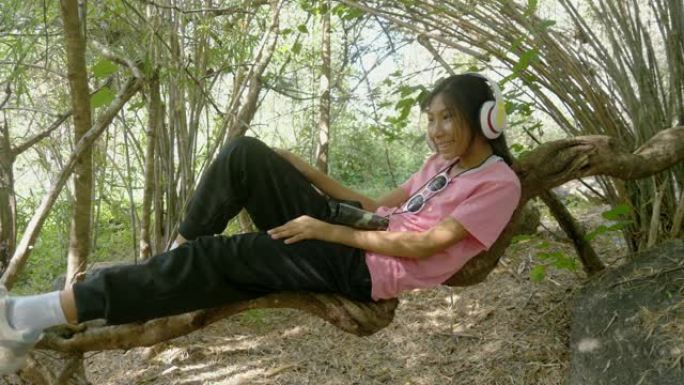 快乐的亚洲女孩在树枝上放松，一边戴着耳机听音乐，生活方式理念。