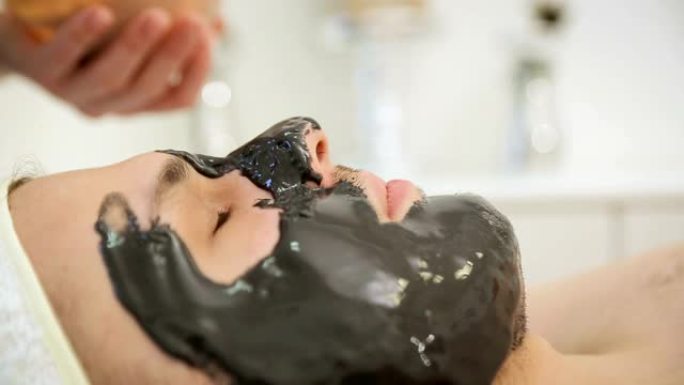 美容师在男人身上戴上黑色面膜，在脸上涂上黑色纸巾的嫩肤化妆品面膜