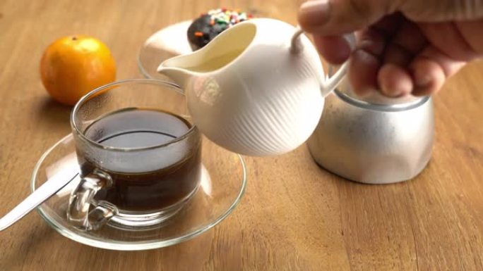 将新鲜牛奶倒入装有成熟橙色和巧克力松饼的木制桌子上的玻璃杯中的黑咖啡中