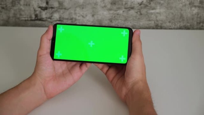 一个坐在白色办公桌前的男人手里拿着手机，屏幕上是绿色的。在智能手机的chromakey屏幕上滑动并点